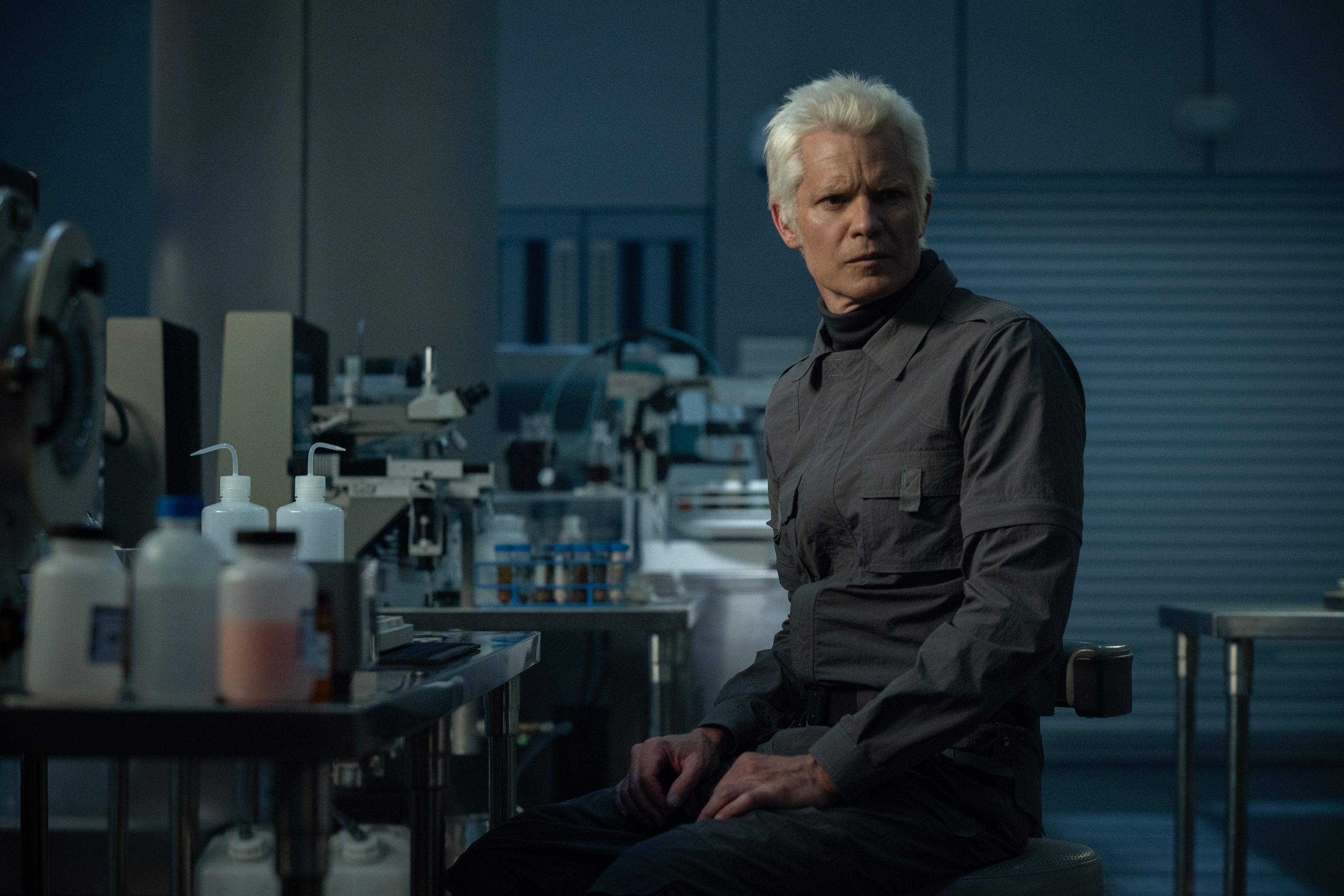Kirsh (Timothy Olyphant, em uma capa de cinza futurista), um andróide de aparência humana, fica em um laboratório escuro em uma cena de Alien Earth
