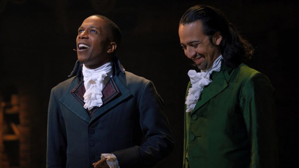 Burr sorrindo e olhando ao lado de Hamilton olhando para baixo rindo em Hamilton