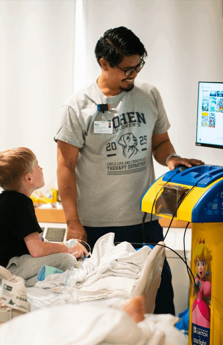 Um pequeno garoto loiro está em uma cama de hospital jogando um videogame, observado por um sorridente assessor de cabelos escuros.