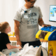Um pequeno garoto loiro está em uma cama de hospital jogando um videogame, observado por um sorridente assessor de cabelos escuros.