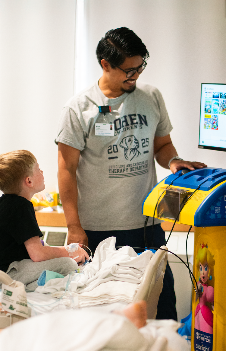 Um pequeno garoto loiro está em uma cama de hospital jogando um videogame, observado por um sorridente assessor de cabelos escuros.
