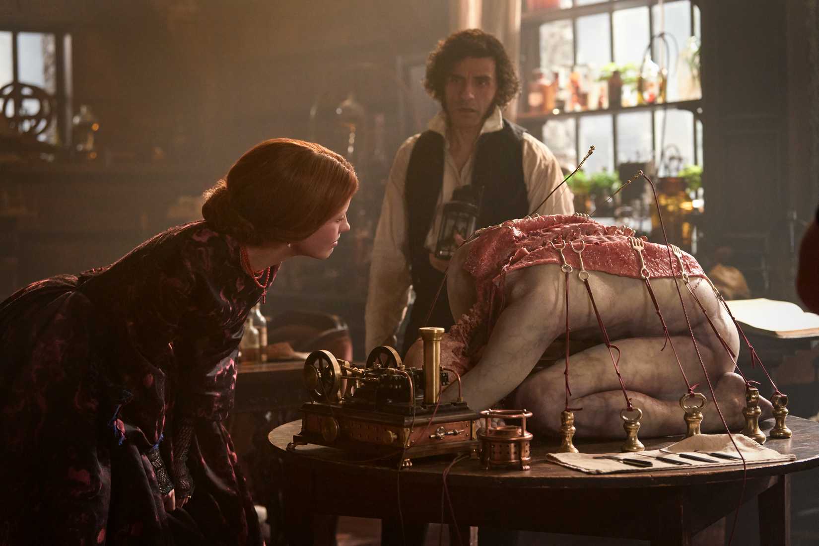 Elizabeth (Mia Goth) examina um cadáver sobre uma mesa enquanto Frankenstein (Oscar Isaac) observa
