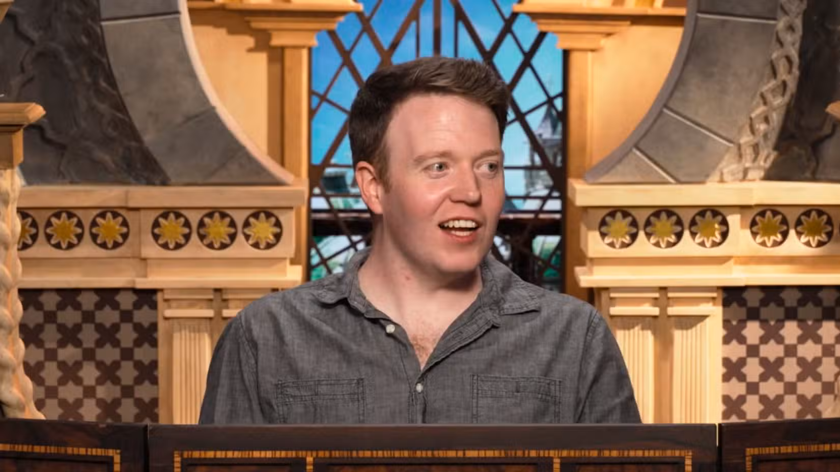 Uma imagem de Brennan Lee Mulligan durante o tempo de espera do Critical Role para o episódio 5. Brennan veste uma camisa cinza de botões abertos e está olhando para o lado com um sorriso.