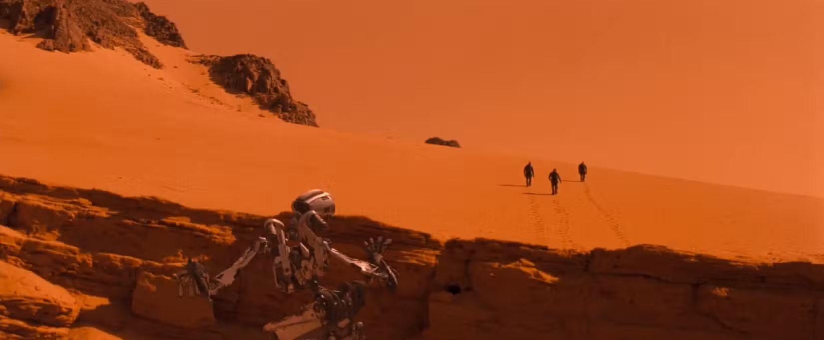 Uma cena do filme Planeta Vermelho retrata uma vasta paisagem marciana em um plano amplo saturado de vermelho-laranja, com três astronautas parecendo minúsculos à distância. 