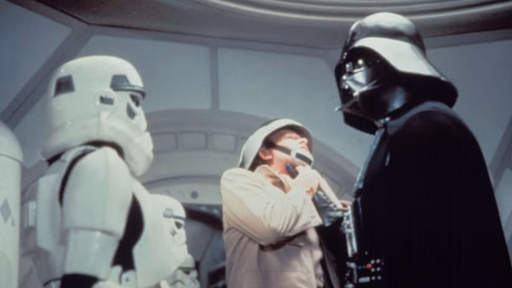 Darth Vader agarra um rebelde pela garganta e o levanta. Um stormtrooper do Império está parado, observando. De Star Wars: Uma Nova Esperança.