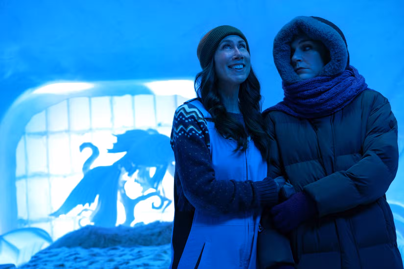 Helen e Carol (Miriam Shor e Rhea Seehorn), embrulhadas em casacos em um quarto iluminado de azul em um hotel de gelo em Pluribus
