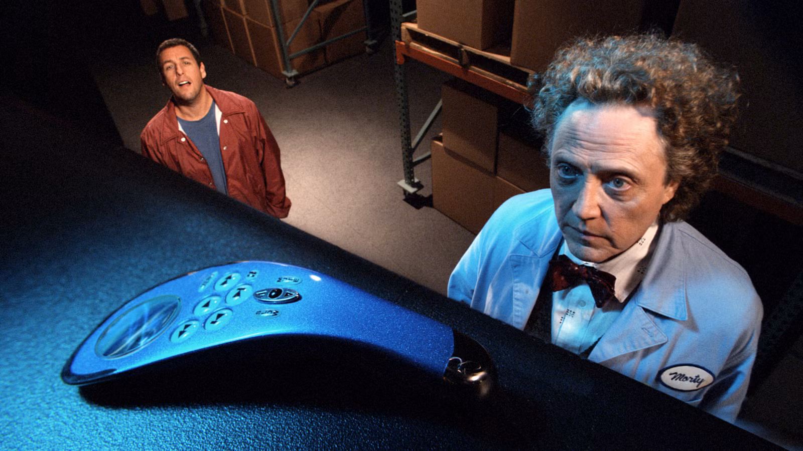Adam Sandler e Christopher Walken olham para um controle remoto pousado de forma suspeita em uma prateleira alta