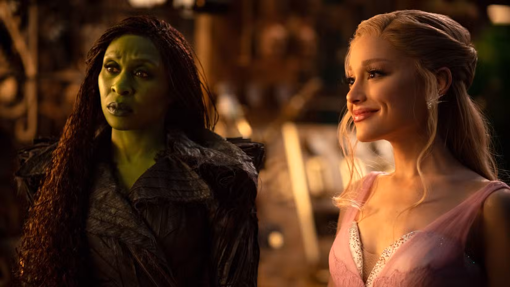 Elphaba Thropp (Cynthia Erivo) e Glinda Upland (Ariana Grande) ficam juntas. Elphaba é verde e usa roupas pretas, inclusive uma capa, e parece desconfiada. Glinda, porém, está vestida de rosa e branco e sorri.
