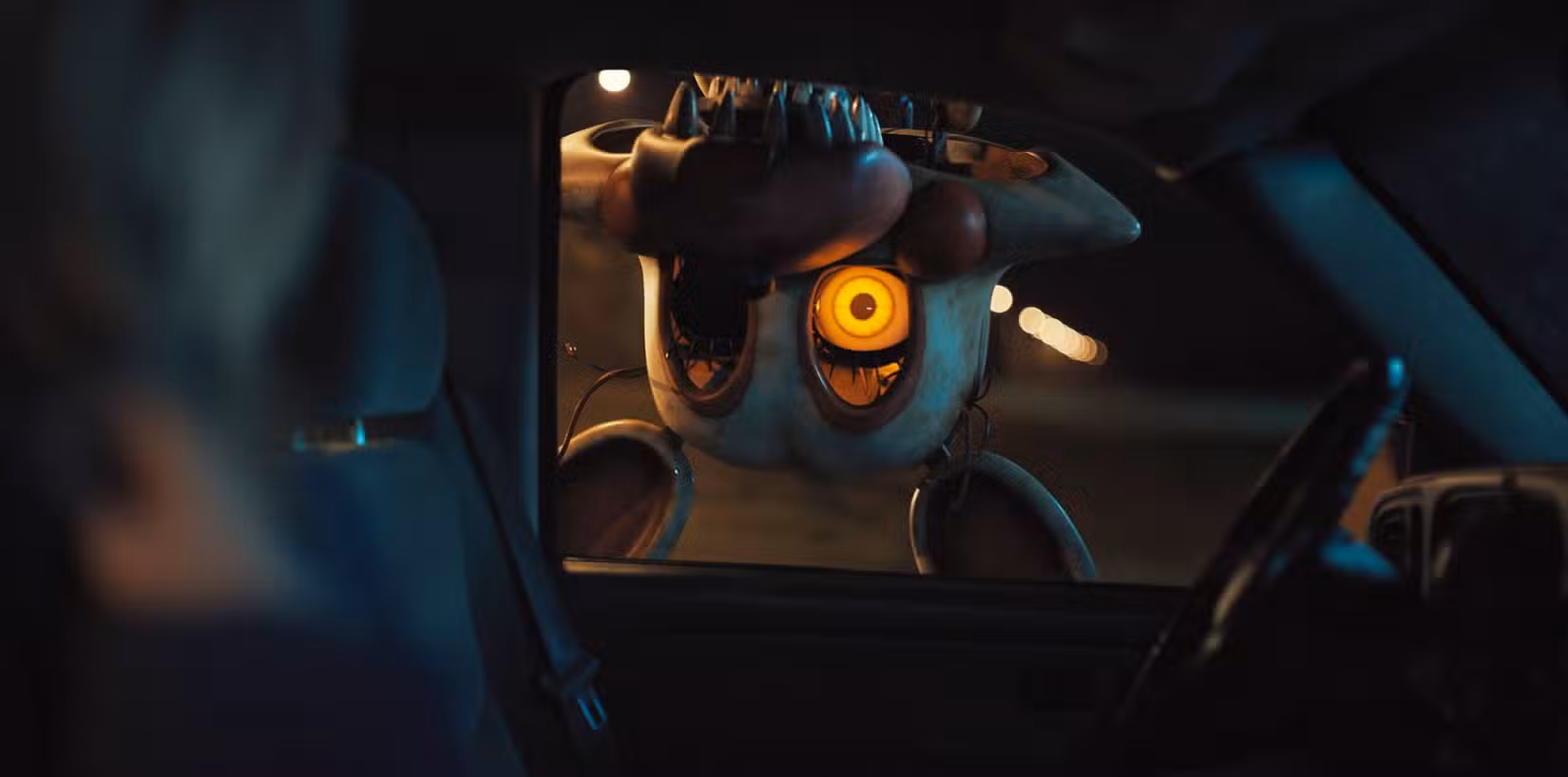 Um animatrônico de cabeça para baixo, sem um olho e dentes afiados e pontiagudos, pendurado do lado de fora da janela de um carro em Five Nights at Freddy's 2