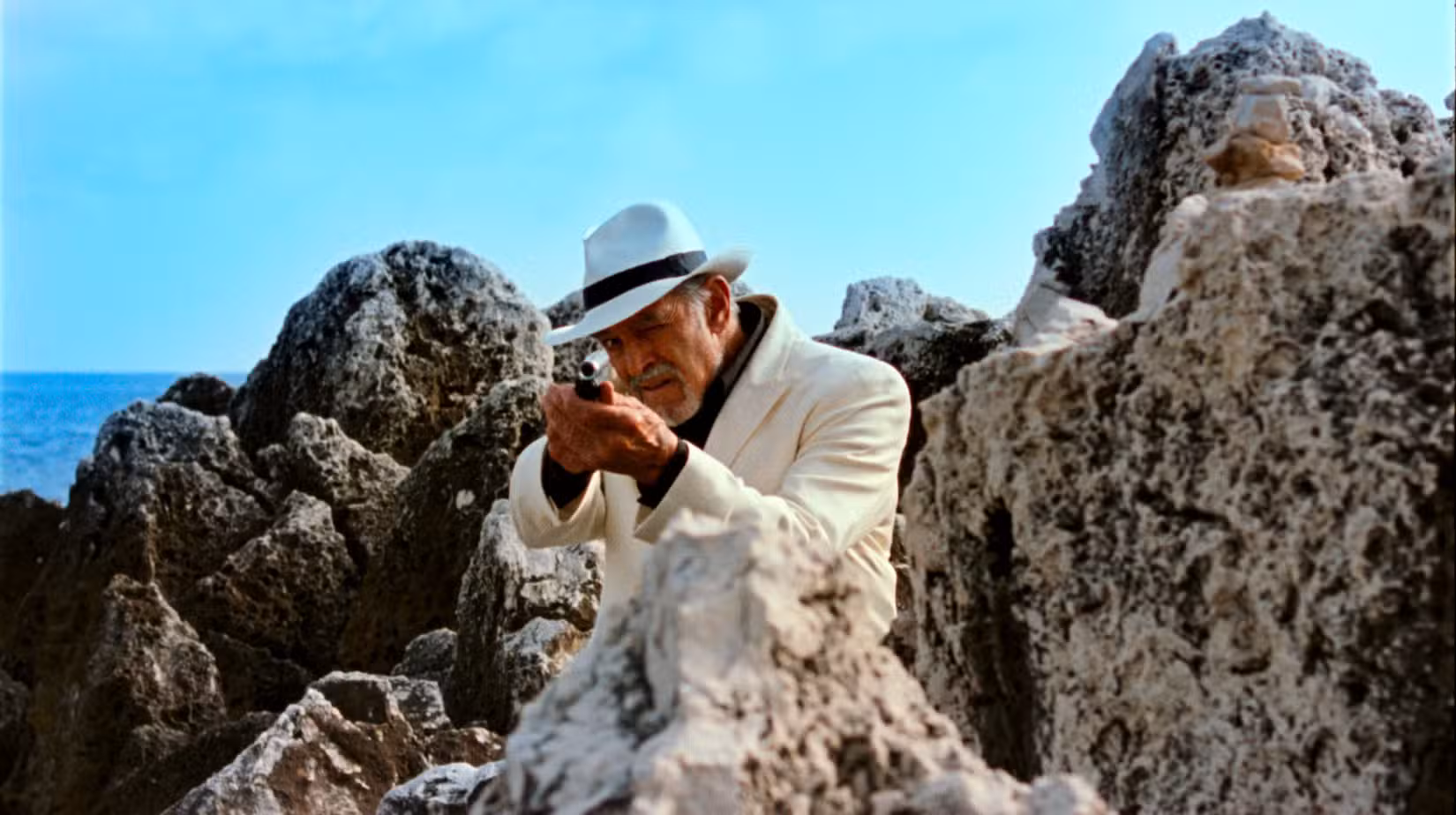Um homem mais velho de terno branco e chapéu de feltro aponta um rifle para a câmera enquanto está parado em meio a enormes rochas porosas na praia em Reflection in a Dead Diamond