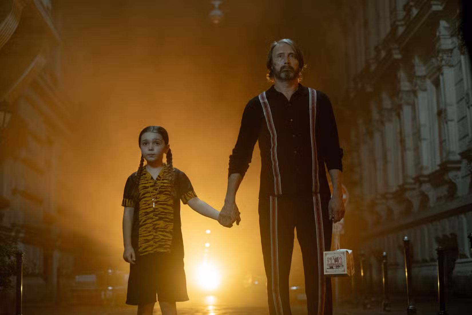 Mads Mikkelsen e Sophie Sloan em Dust Bunny, de Bryan Fuller. Sloan usa uma camisa e shorts listrados de tigre, enquanto Mikkelsen usa uma camisa e calças finamente decoradas. Uma luz brilhante brilha no beco atrás deles.
