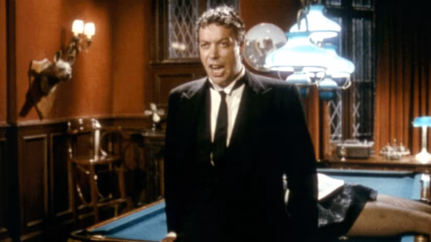 O mordomo Wadsworth (Tim Curry) explica o mistério do assassinato enquanto está em frente a uma mesa de sinuca com um cadáver em Clue