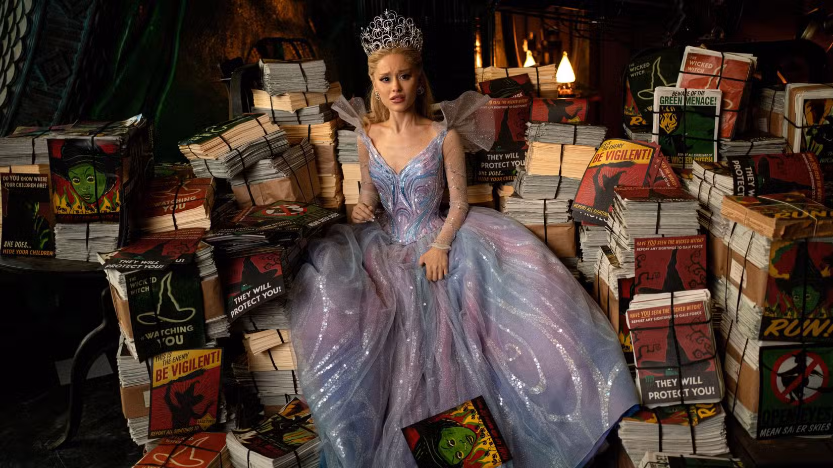 Glinda (Ariana Grande) está sentada em cima de uma pilha de diferentes pôsteres da Bruxa Malvada do Oeste. Ela parece enojada.