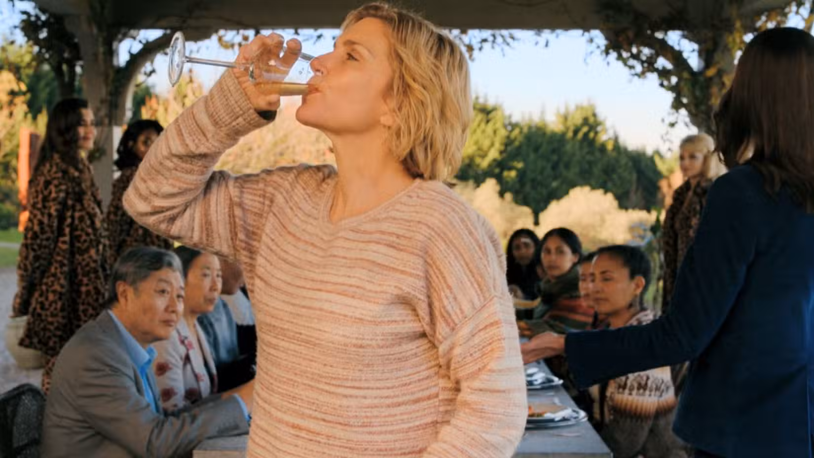 Uma captura de tela do episódio 2 de Pluribus apresentando Rhea Seehorn como Carol Sturka. Ela bebe de uma taça de vinho, cercada por seus colegas imunes e um bando de mentes coletivas.