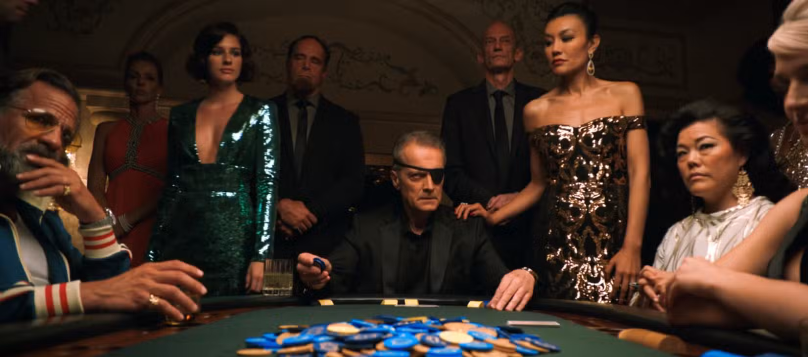 Um homem de aparência severa e tapa-olho está sentado em uma mesa de pôquer em Las Vegas, cercado por espectadores sérios, no episódio 6 de Pluribus