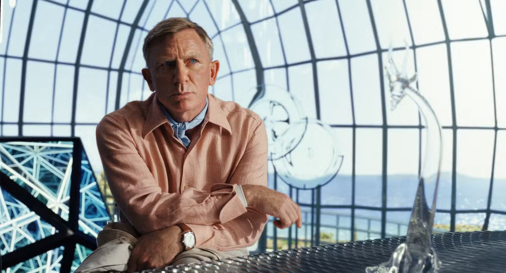 Daniel Craig como Benoit Blanc em Glass Onion, parecendo pensativo em uma camisa de linho rosa e gravata azul dentro de uma cúpula de vidro multifacetada
