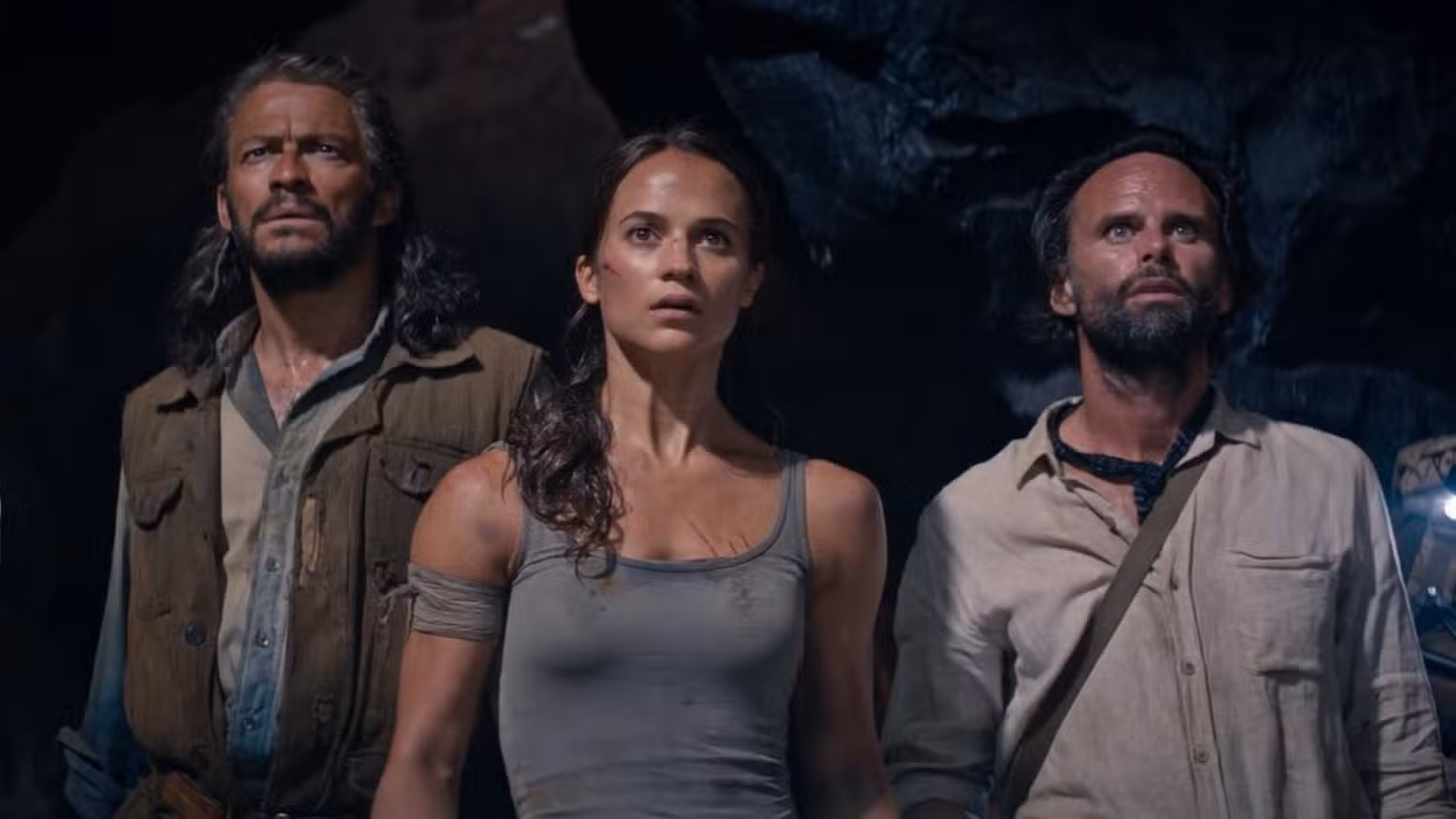 Dominic West, Alicia Vikander e Walton Goggins, alinhados em fila, olham maravilhados para, o que mais, uma tumba particularmente elaborada (fora da tela), em uma cena da versão 2018 de Tomb Raider. 