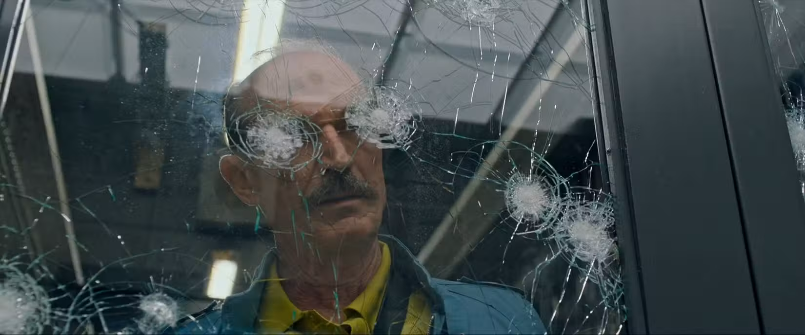 Toby Huss, interpretando um assassino maníaco, olha através de uma janela crivada de balas, com duas rachaduras de bala alinhadas para cobrir seus olhos, em uma cena de Copshop de Joe Carnahan (2021)