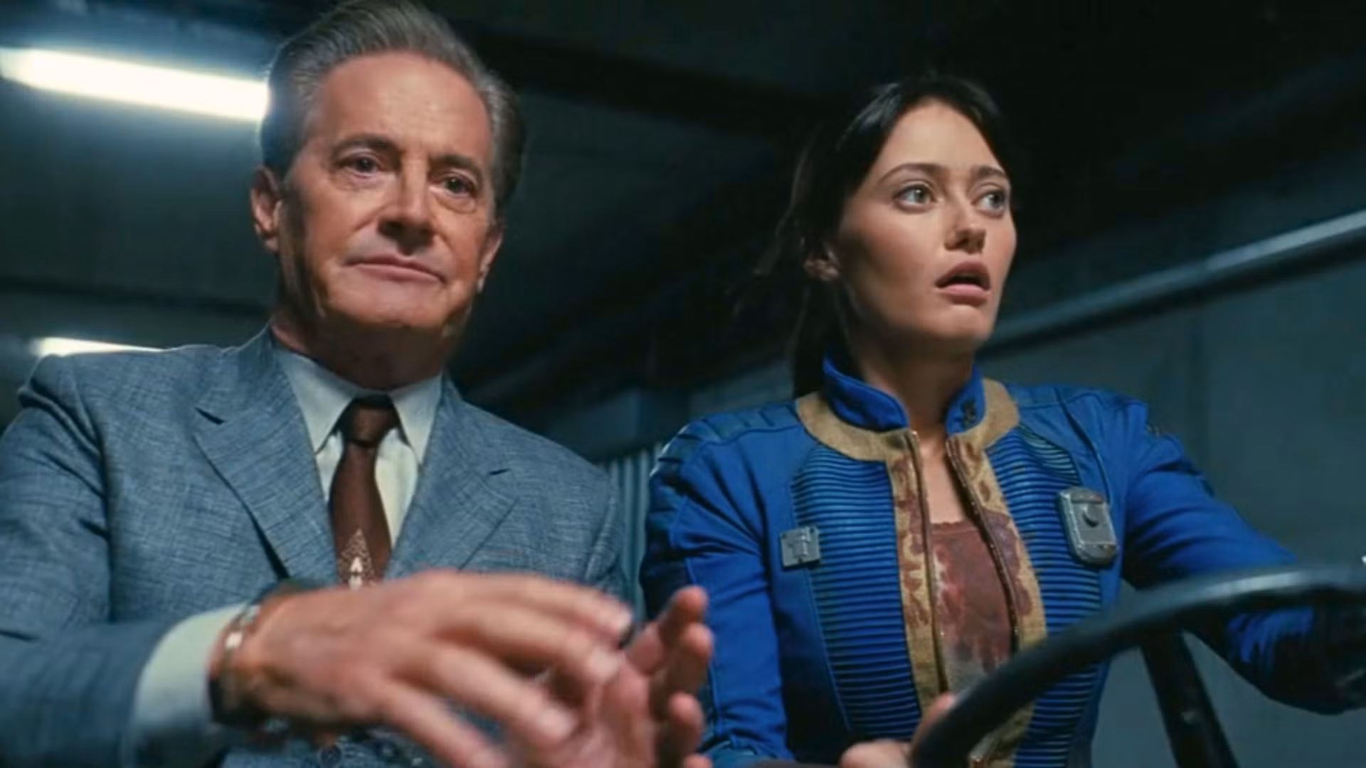 Uma imagem da segunda temporada de Fallout do Prime Video. Ela mostra Hank, um senhor mais velho em um terno elegante, tentando tirar o volante de Lucy, sua filha. Ela usa um traje de salto azul.