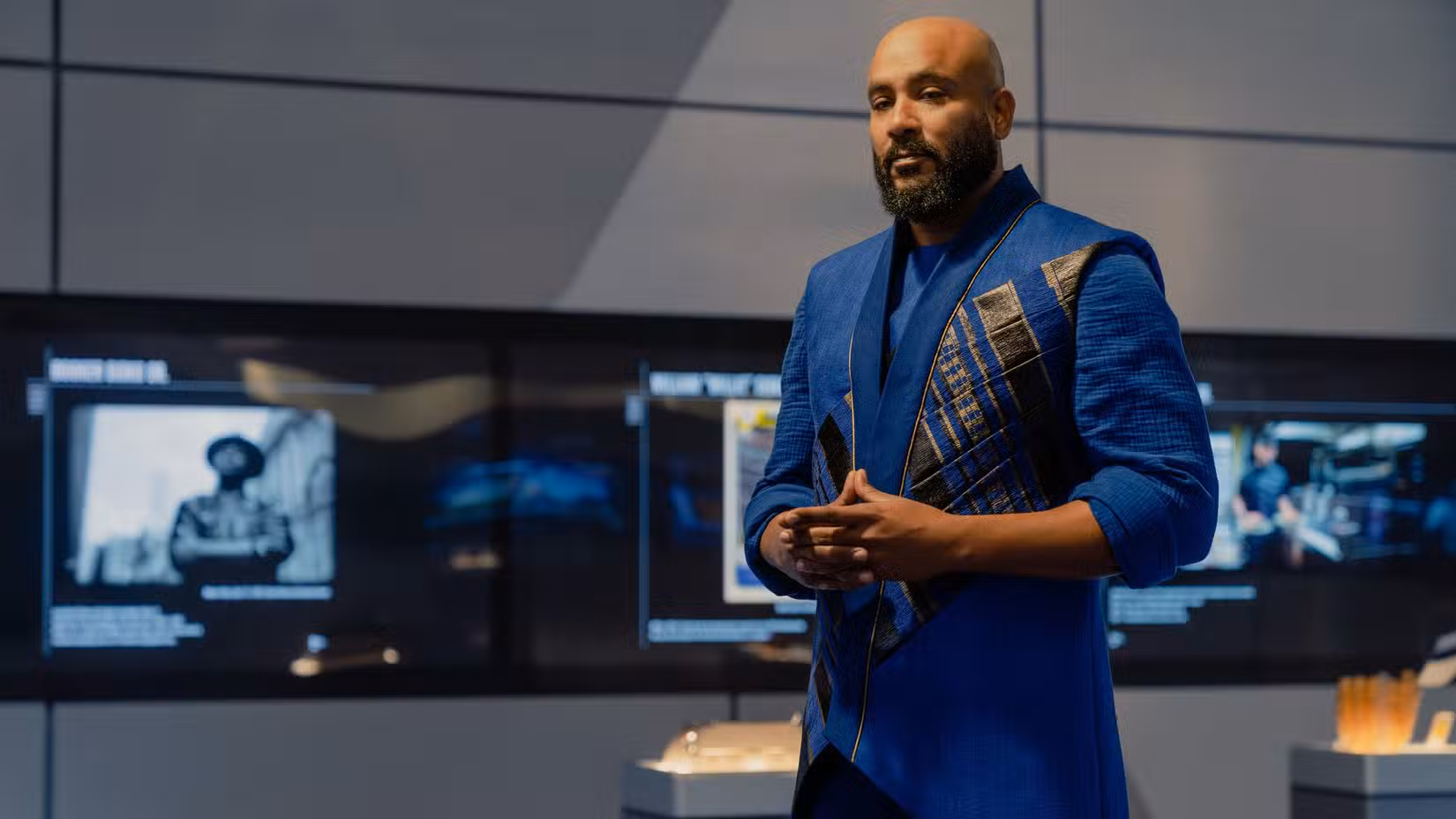 Jake Sisko (Cirroc Lofton), um homem de meia-idade com barba e uma jaqueta azul e dourada, está no museu Sisko na Academia da Frota Estelar parecendo contemplativo