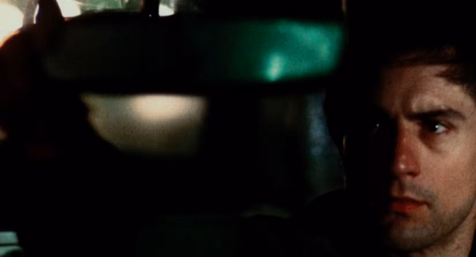 O rosto de Travis Bickle (Robert De Niro) é visível no canto inferior direito de uma visão distorcida e embaçada de seu táxi e do mundo exterior, enquanto ele olha pelo espelho retrovisor para algo que o público não consegue ver.