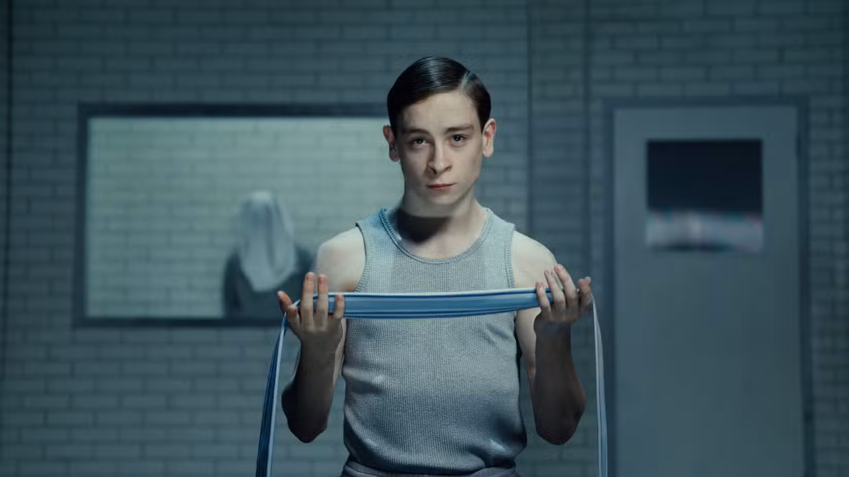 Louis McCartney em Stranger Things: The First Shadow, parado em uma sala institucional de azulejos cinza com uma figura mascarada atrás dele