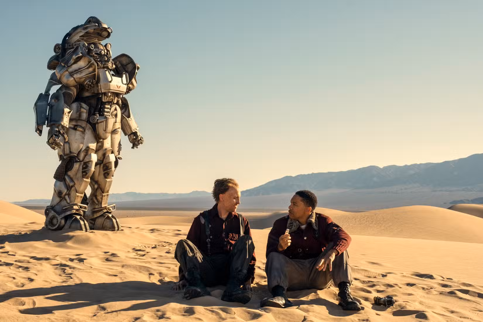 Johnny Pemberton (Thaddeus) e Aaron Moten (Maximus) sentam-se na areia com uma armadura de poder atrás deles na 2ª temporada de Fallout