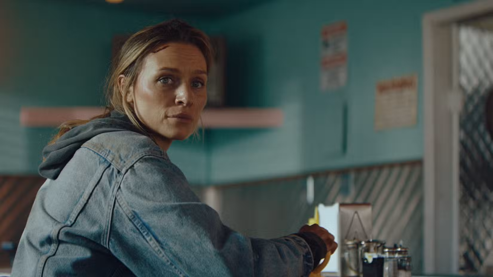 Irene (Michaela McManus), uma mulher de meia-idade vestindo uma jaqueta jeans com um corte na cabeça, olha por cima do ombro enquanto está sentada em uma lanchonete em Redux Redux.