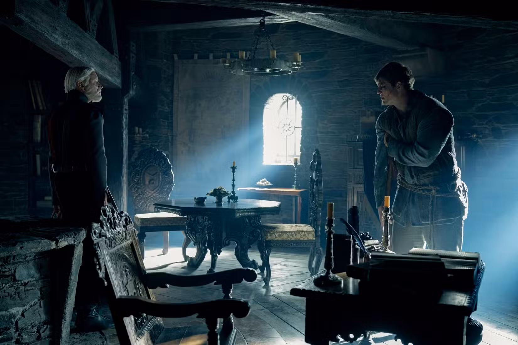 Dois homens, um mais velho e vestido de preto, um mais jovem, gravemente machucado e apoiado em uma muleta, encaram-se em uma sala medieval muito escura, com luz entrando por uma janela central em Um Cavaleiro dos Sete Reinos