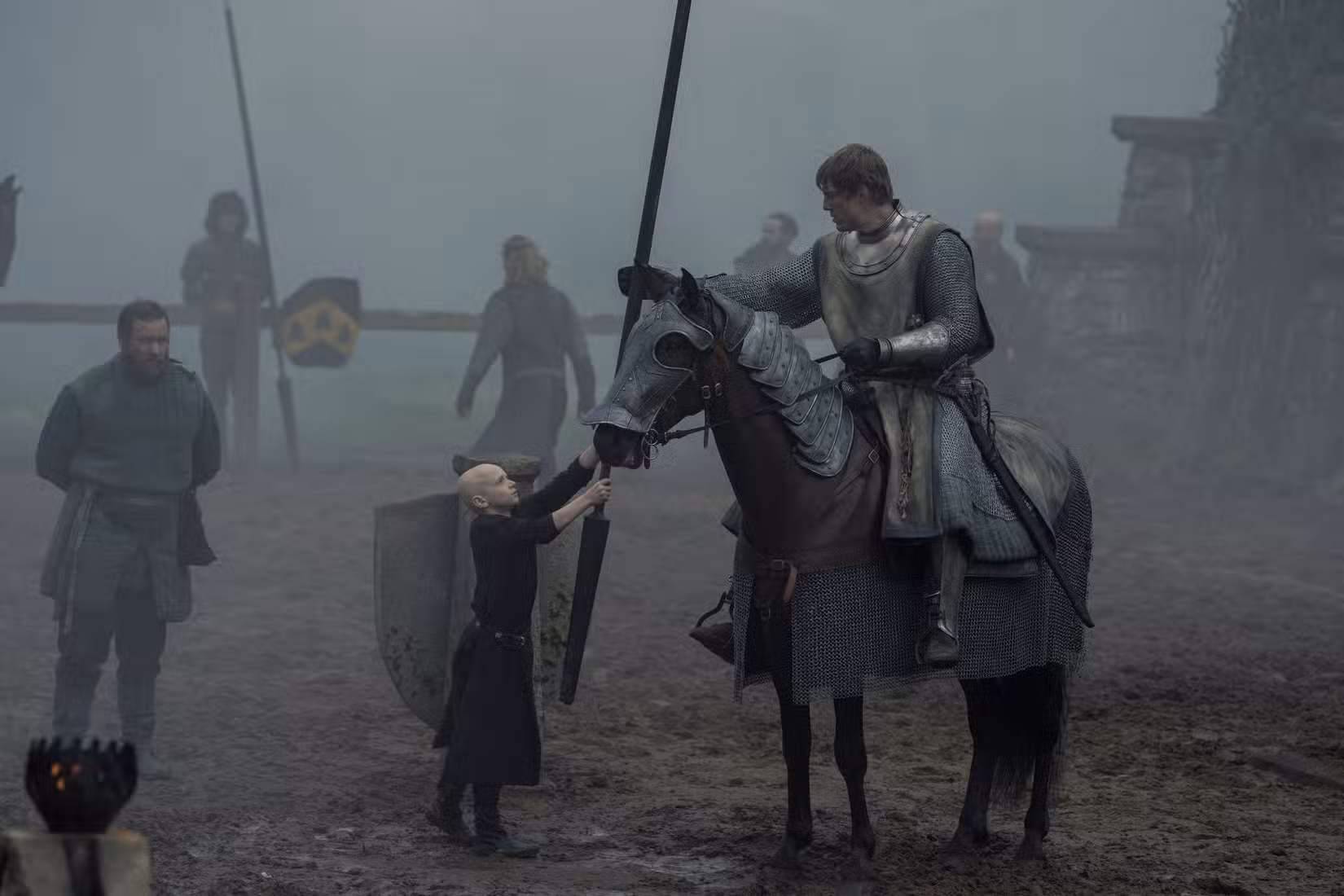 Um cavaleiro a cavalo (Peter Claffey) aceita uma lança de um garotinho careca (Dexter Sol Ansell) em uma arena de justas enevoada em O Cavaleiro dos Sete Reinos