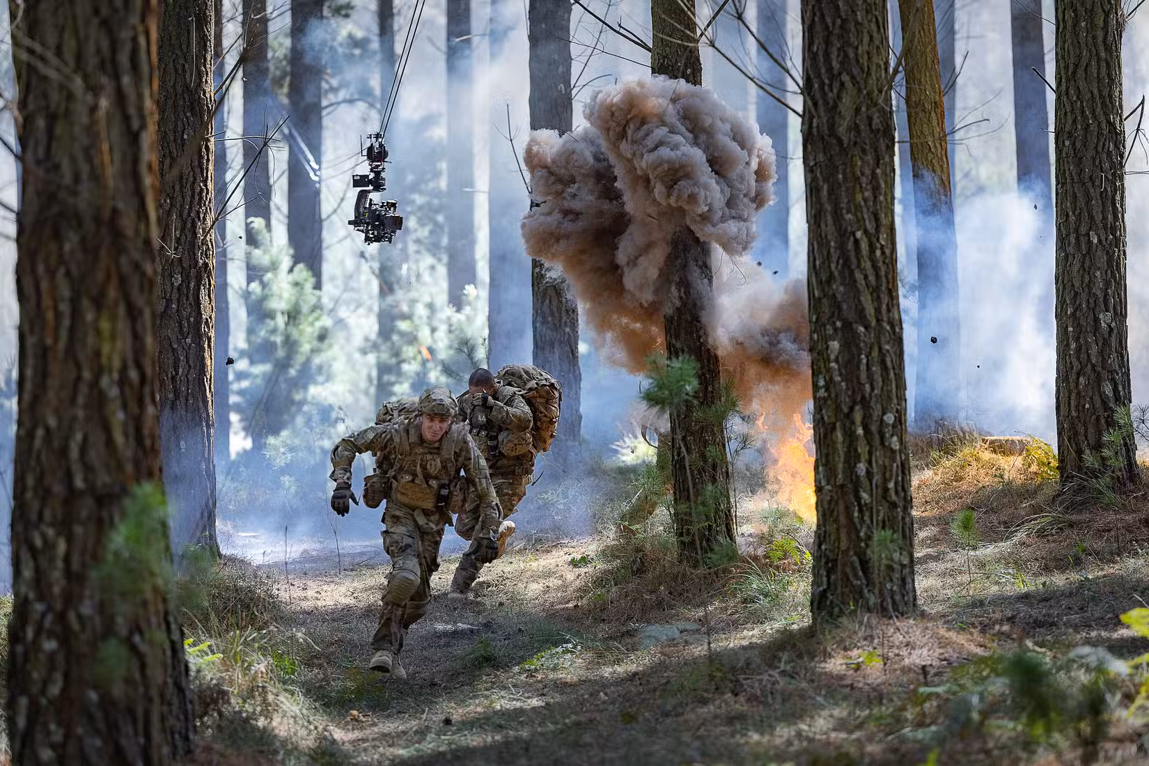 Atores vestidos como estagiários do Exército correm por entre árvores enquanto explosões são desencadeadas e uma câmera paira sobre fios no set de War Machine