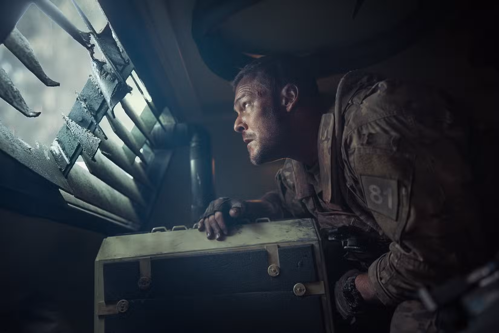 O sargento 81 do exército (Alan Ritchson), parecendo nervoso, fica curvado dentro de um caminhão blindado e espia através de uma janela com persianas em War Machine.