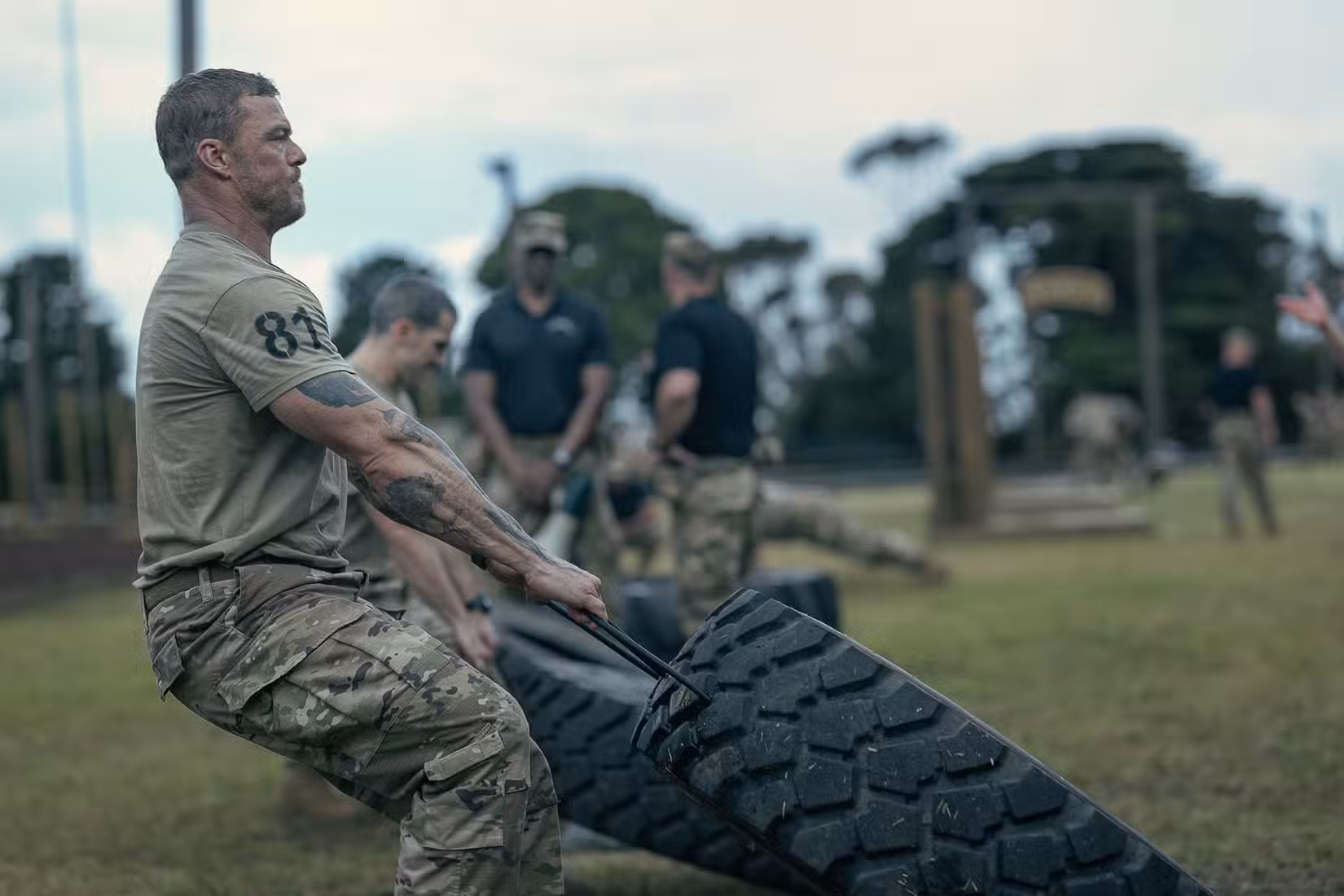 81 (Alan Ritchson), um sargento do Exército com calças camufladas e camiseta, cerra a mandíbula enquanto arrasta um pneu enorme ao lado de uma fila de outros candidatos a Rangers fazendo o mesmo em War Machine