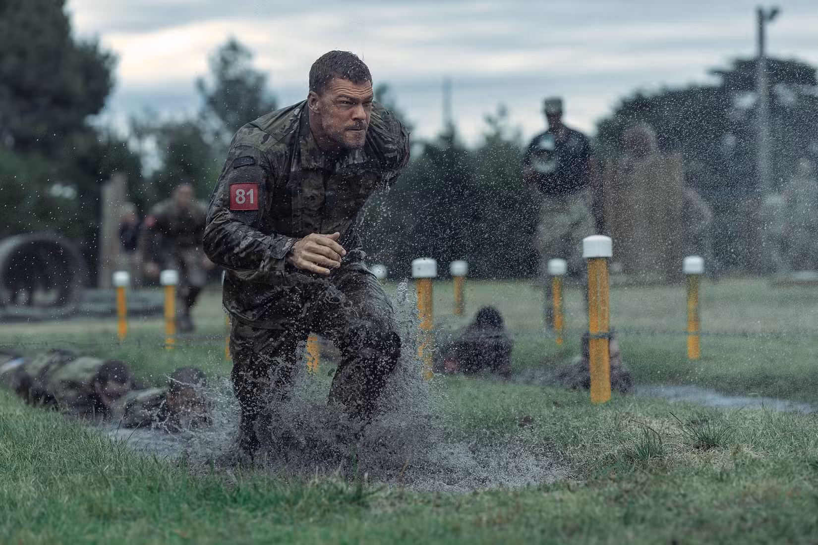 O sargento 81 do exército (Alan Ritchson), encharcado de uniforme e número de identificação vermelho, corre por um campo encharcado em meio a postes amarelos de sinalização de pista em War Machine