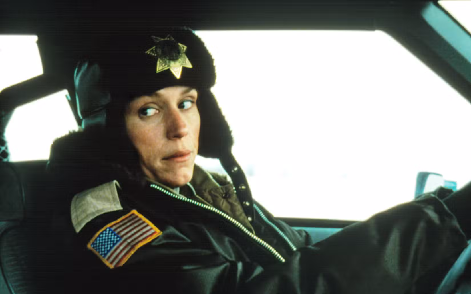 Marge Gunderson (Frances McDormand) olha por cima do ombro enquanto dirige seu vagabundo em Fargo, vestindo uma jaqueta pesada e um chapéu de pele