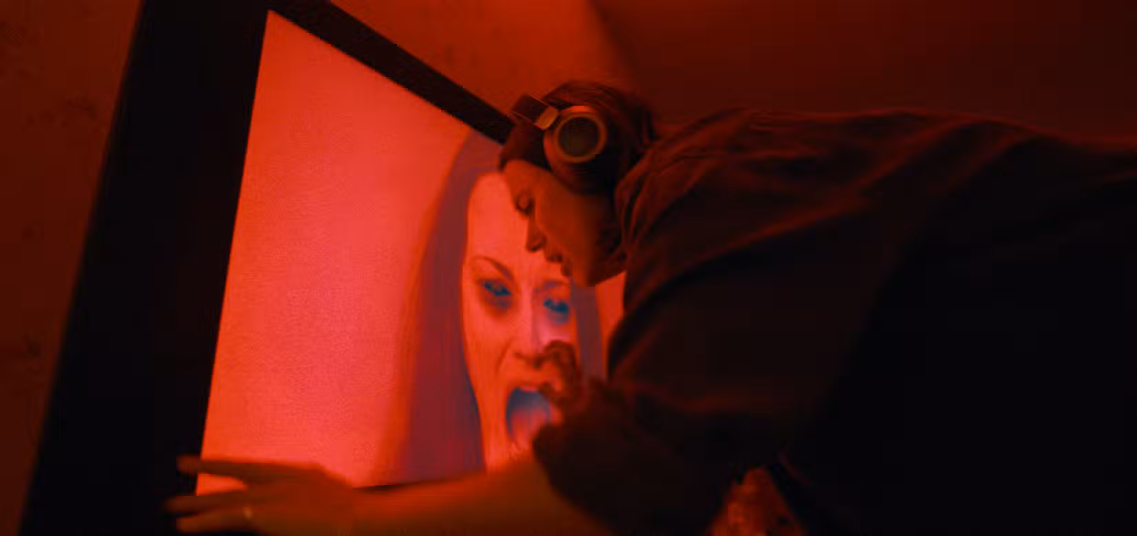 Evy (Nina Kiri), uma jovem com cabelos escuros e fones de ouvido, se inclina sobre uma TV de tela plana exibindo uma imagem iluminada em vermelho de uma mulher gritando com olhos escurecidos em Undertone