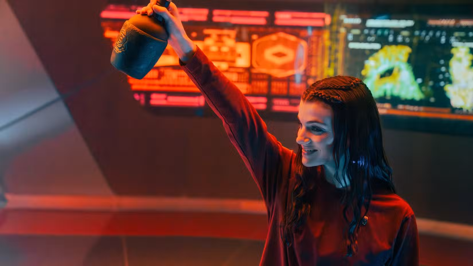 Tarima Sadal (Zoë Steiner) segura uma jarra com uma bebida fortemente alcoólica em frente a um painel de telas do USS Athena