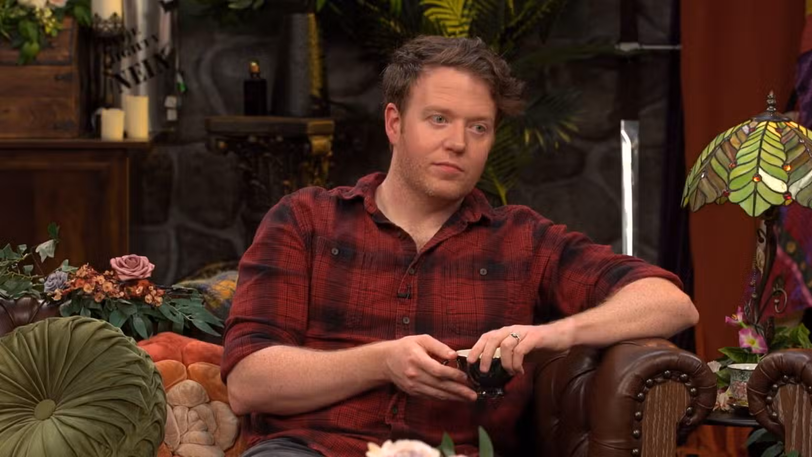 Uma imagem de Brennan Lee Mulligan sentada em um sofá no set de Critical Role. Ele veste uma camisa xadrez vermelha e preta e tem uma xícara de chá na mão.