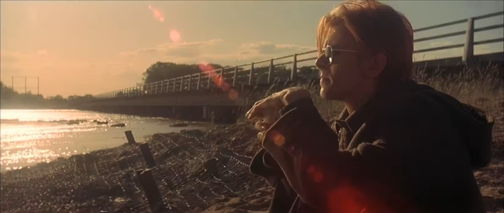 Um alienígena em forma humana (interpretado por David Bowie, é claro) está sentado na margem de um lago, usando óculos escuros e parecendo contemplativo enquanto o sol brilha atrás dele em uma cena de O Homem que Caiu na Terra (1976).