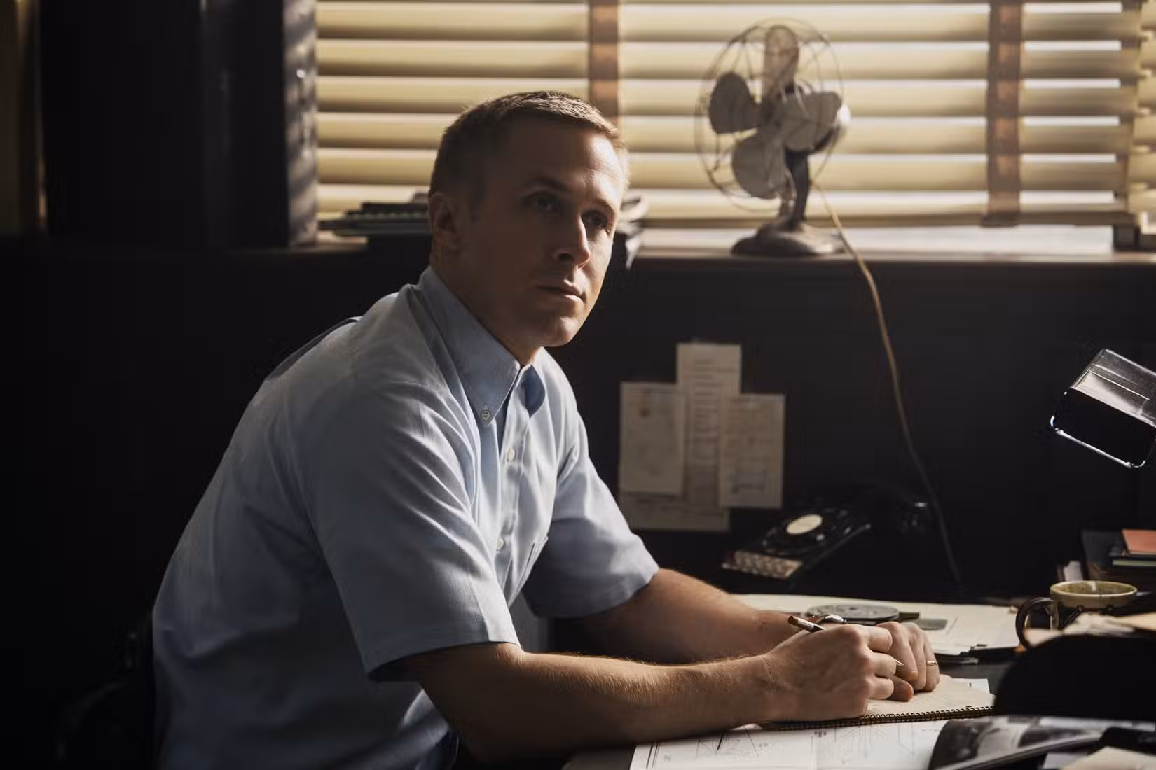 Neil Armstrong (Ryan Gosling), de corte militar e aparência séria, ocupa uma mesa em uma cena de First Man (2018). 