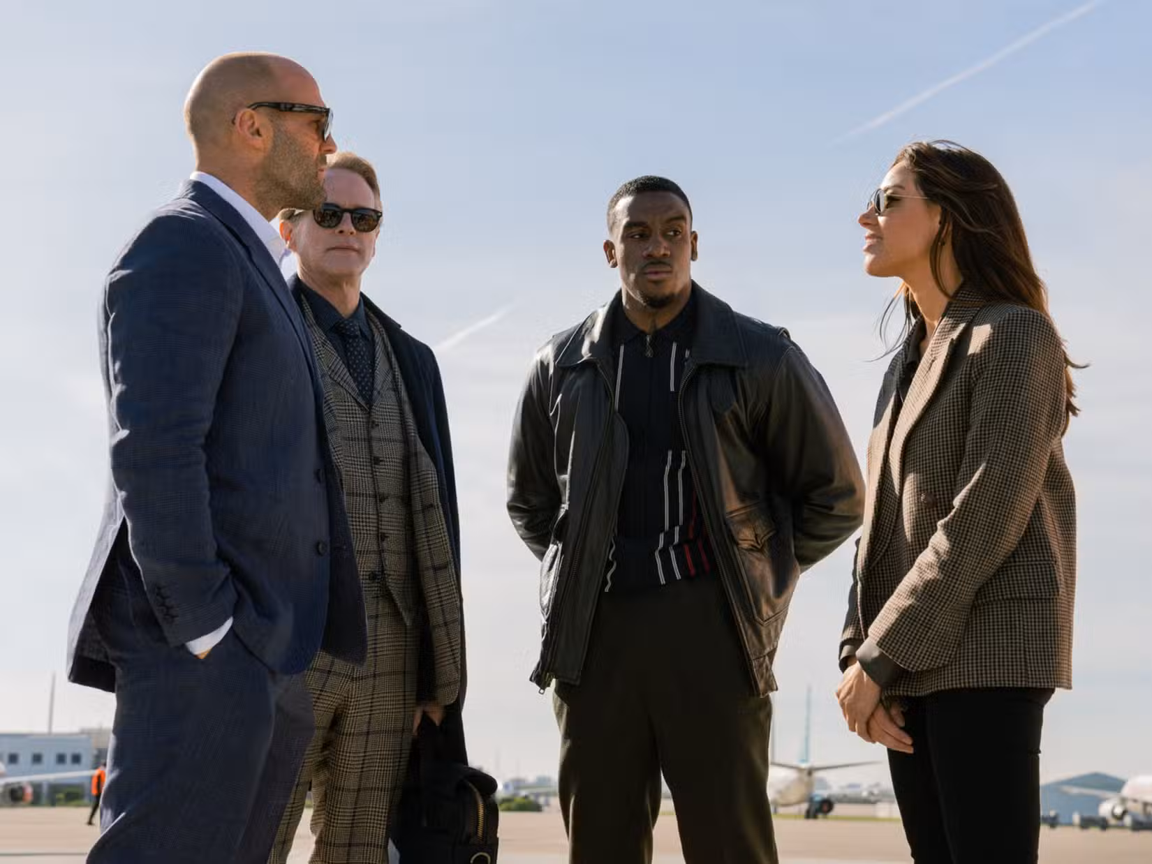 Jason Statham, Cary Elwes, Bugzy Malone e Aubrey Plaza conversam juntos na pista de um avião em uma cena da Operação Fortune: Ruse de Guerre.