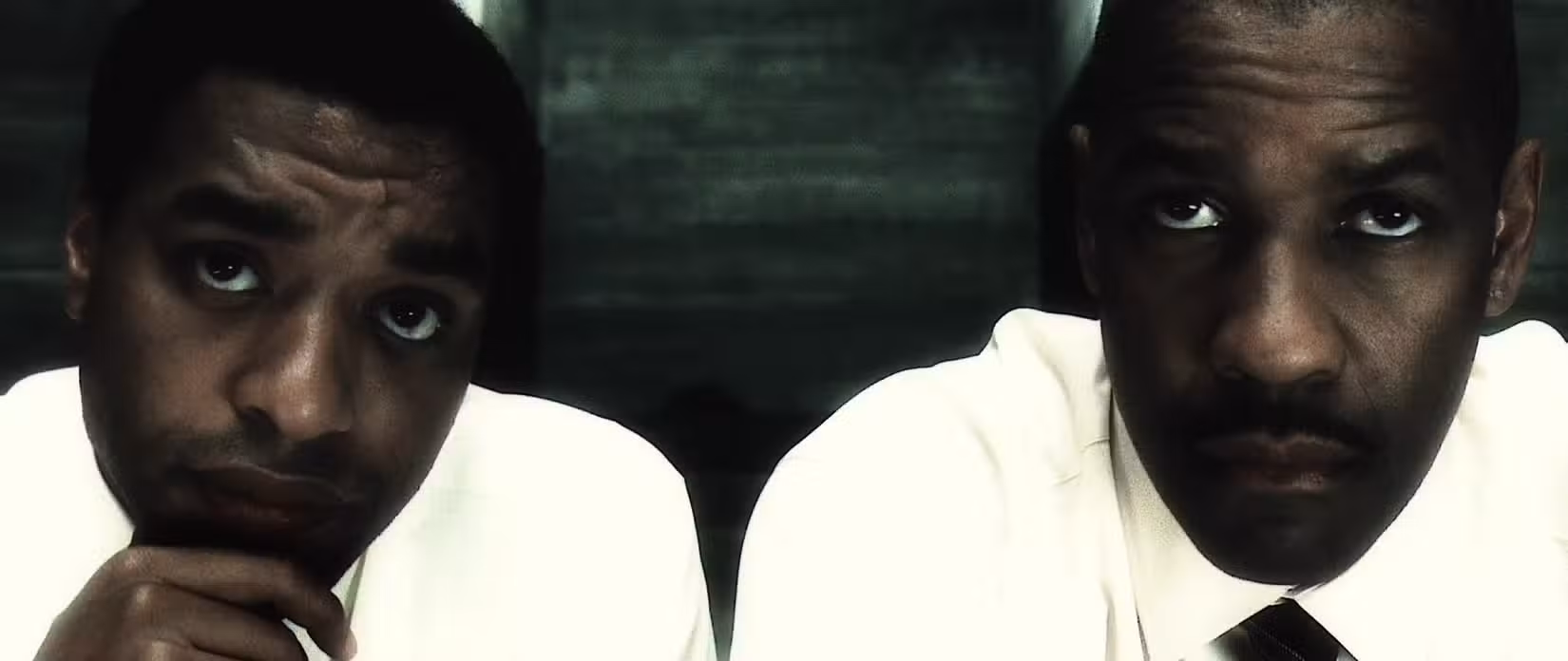 Denzel Washington e Chiwetel Ejiofor sentam-se lado a lado em close-up em uma cena de Inside Man que usa um efeito descolorido de alto contraste no filme para tornar suas jaquetas brancas especialmente brilhantes.  