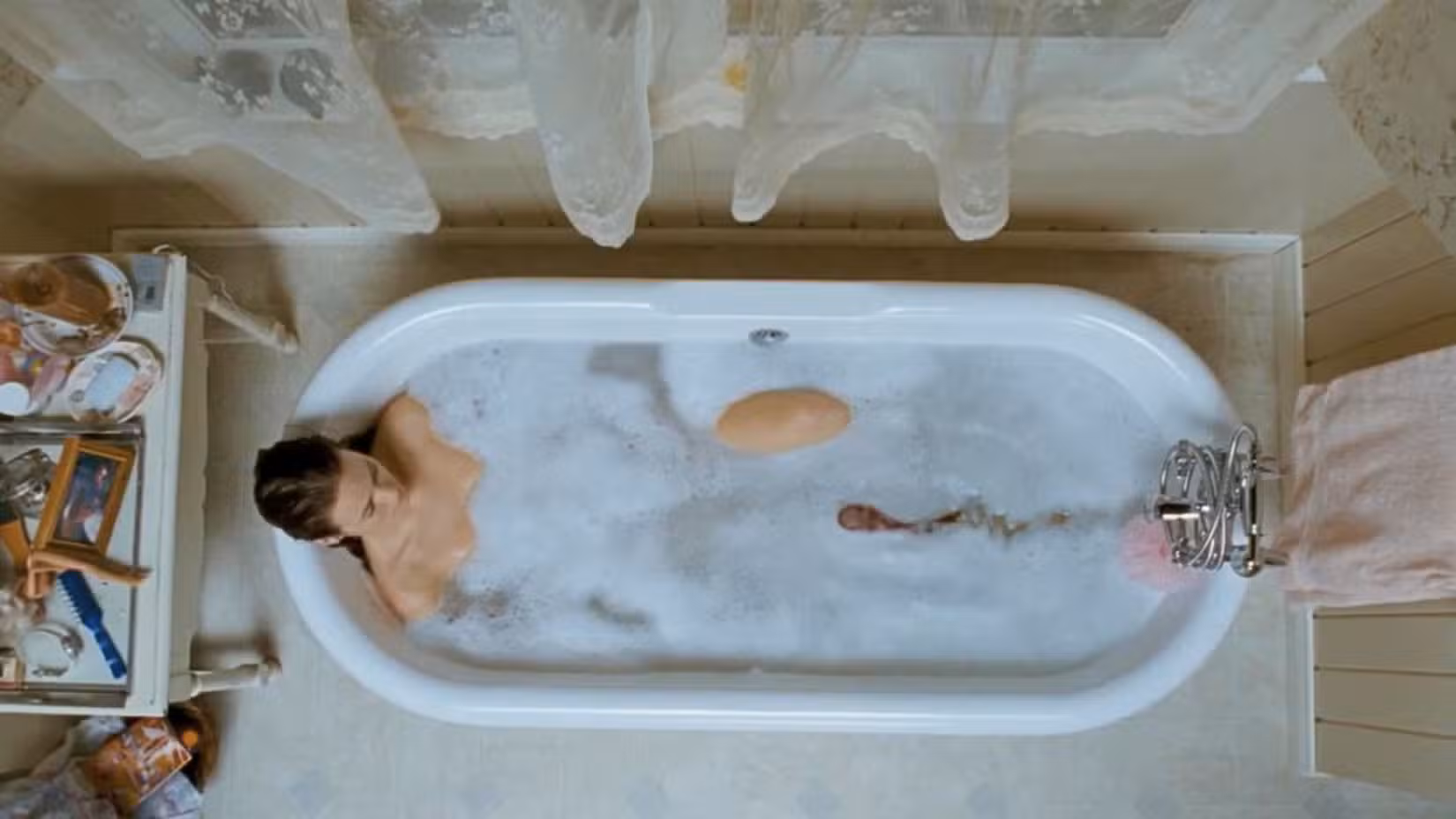 Uma mulher está deitada na banheira com uma criatura parecida com uma lesma vagando pelo sabão e pela água com sabão em sua direção.