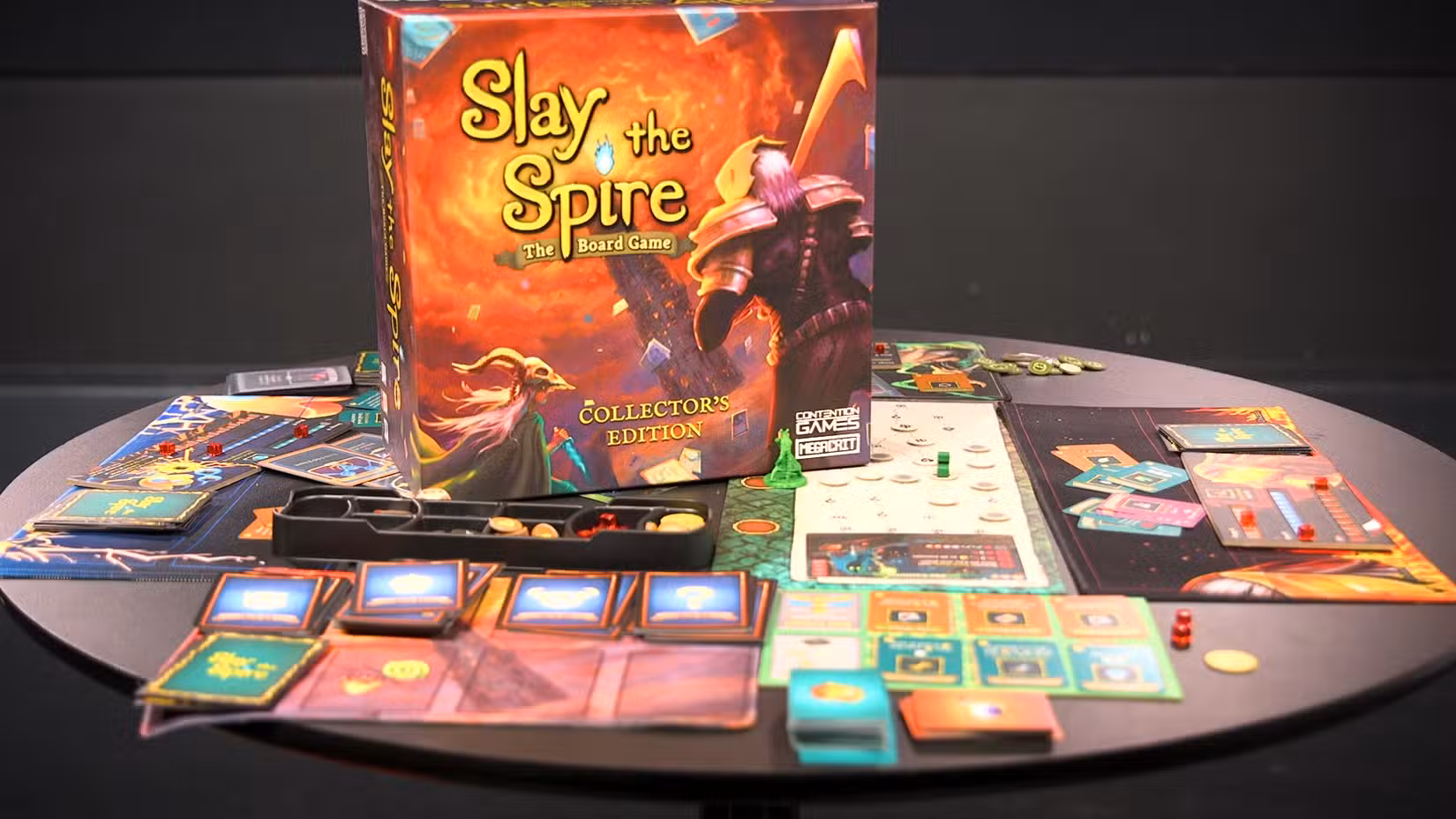 Uma mesa coberta de cartas e tapetes de jogador do jogo de tabuleiro Slay the Spire, com a caixa do jogo bem no meio.