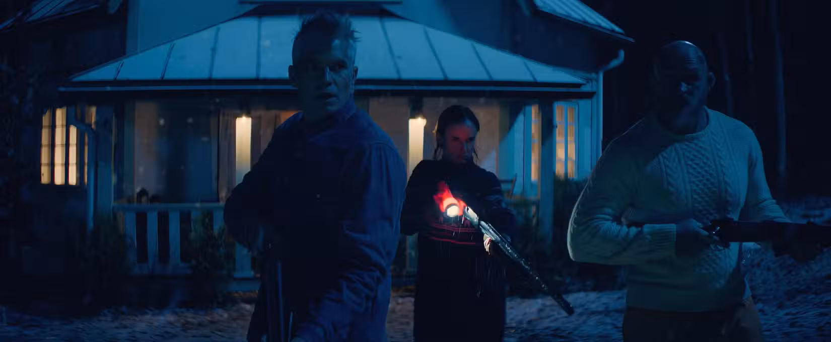 Timothy Olyphant, Juliette Lewis e Keith Jardine, todos segurando armas, cruzam em frente a uma casa iluminada à noite em Over Your Dead Body