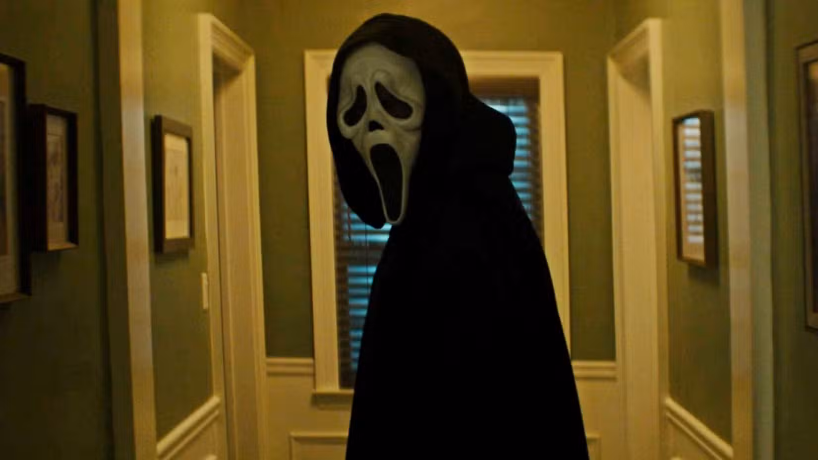 Uma figura com capuz e máscara Ghostface olha para um corredor, emoldurado no centro, em uma cena de Pânico 7