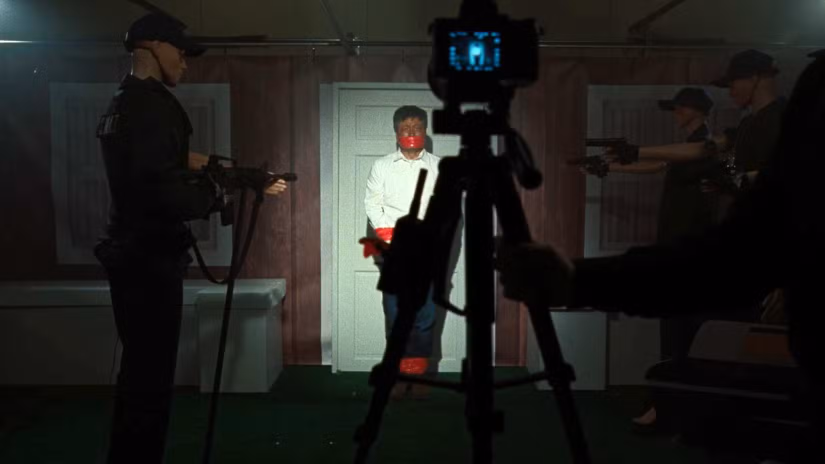 Em um porão mal iluminado com uma câmera de vídeo montada para gravá-lo, manequins são montados para representar um homem do lado de fora de uma porta cercado por policiais armados, em cena de Faces of Death (2026) recriando uma cena do filme de 1978 de mesmo nome.