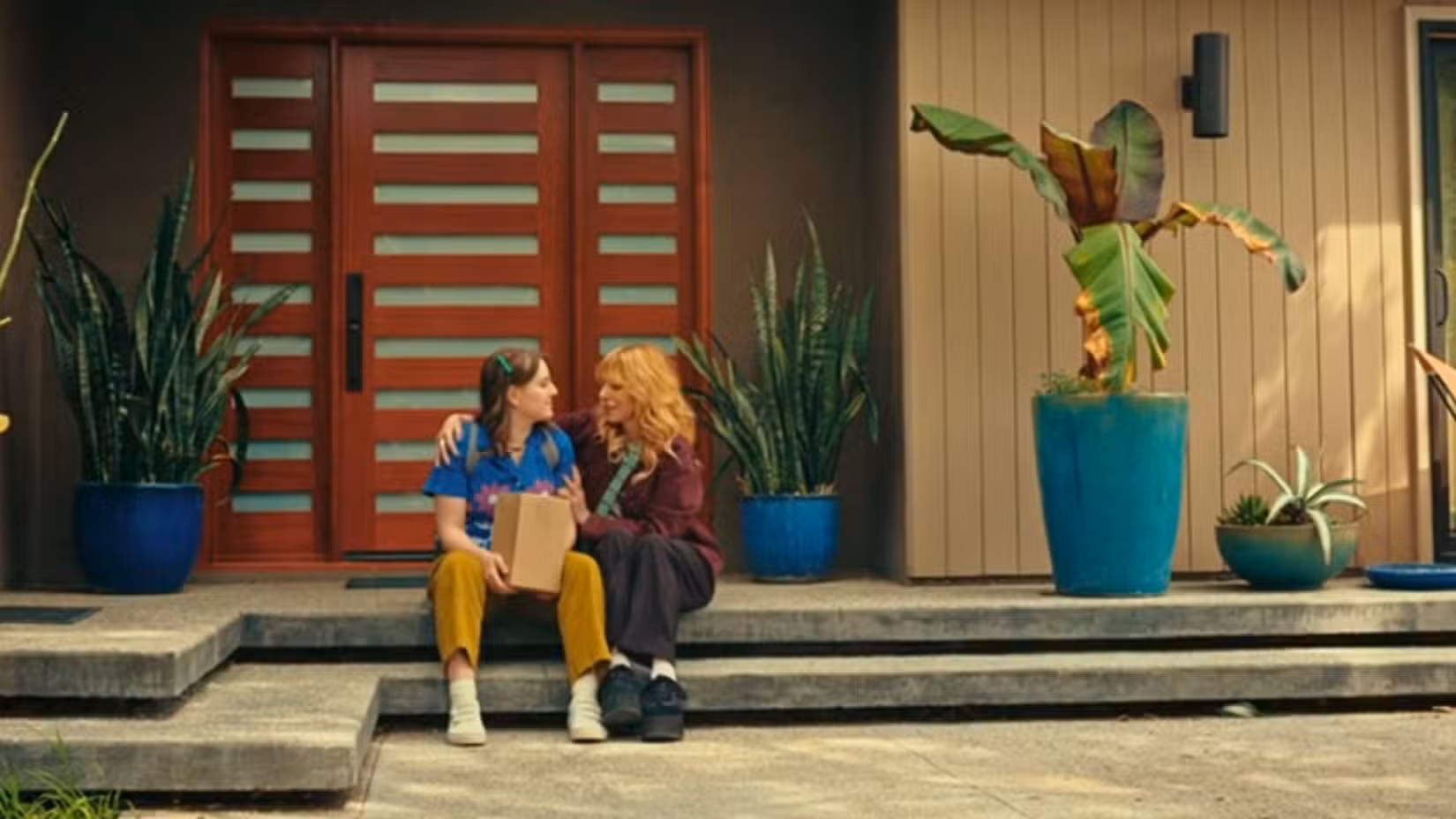 Uma imagem de D(e)ad. Mostra Tillie, uma jovem, de camisa azul e calça amarela, sentada ao lado de uma mulher loira vestida de vermelho e preto. Eles estão abraçados.