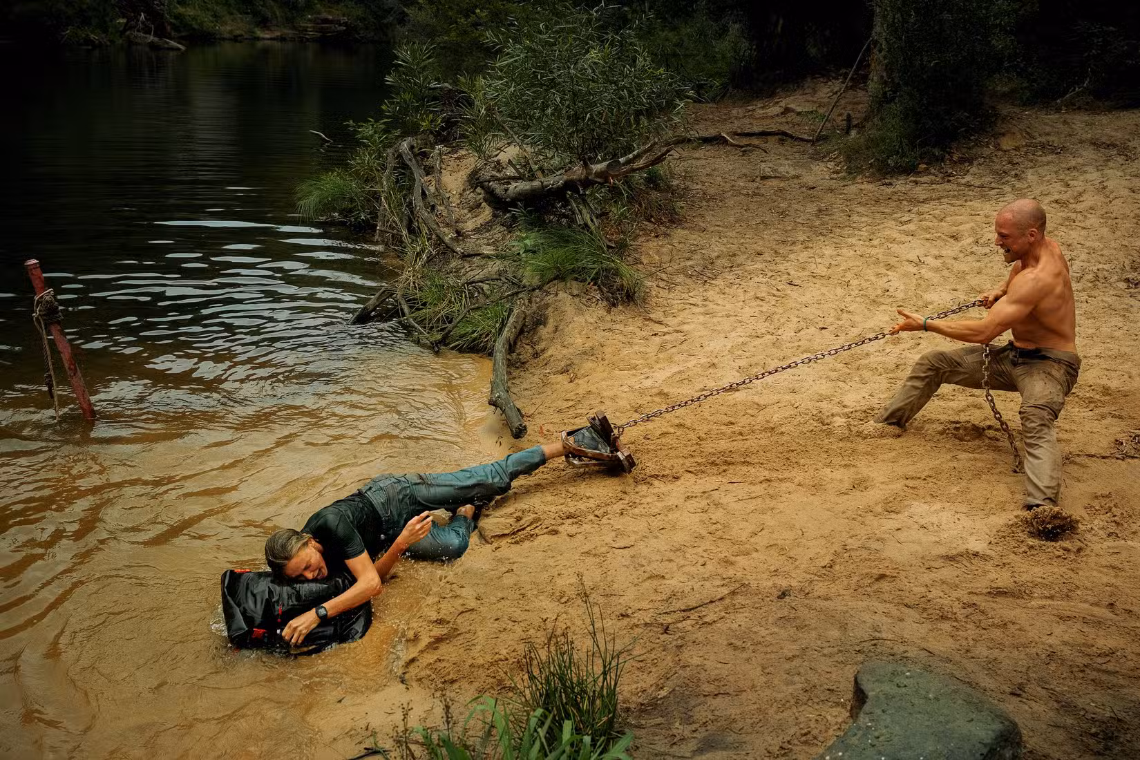 Taron Egerton transporta Charlize Theron por uma praia arenosa por uma corrente presa a uma armadilha para ursos em sua perna em Apex