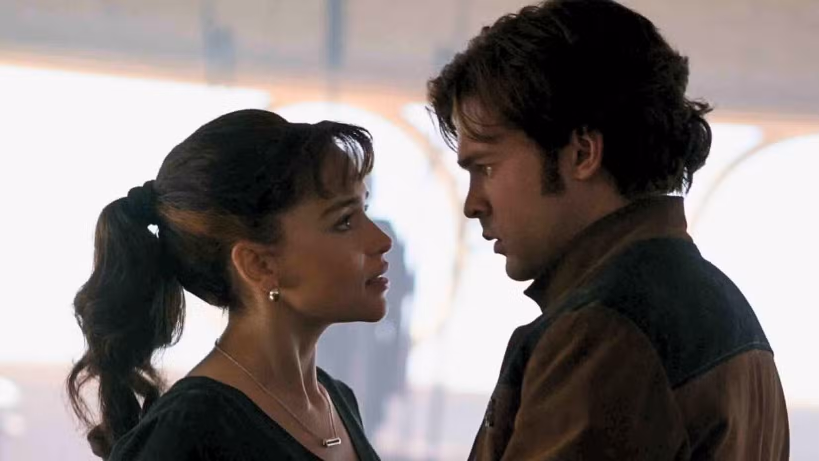 Han e Qi'ra de Star Wars: Solo. Qi'ra tem cabelo castanho preso em uma ponta ondulada com franja na testa. Ela usa roupas pretas e olha preocupada para Han. Han está com uma jaqueta marrom e bege e retorna sua expressão preocupada.